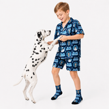 A young boy wearing a blue and white printed pajama set with matching socks is standing next to a Dalmatian dog, reaching out to pet it, against a plain white studio background.