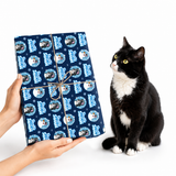 A black and white cat sits next to a person holding a wrapped gift box. The gift box is wrapped in navy blue wrapping paper with a repeating pattern of cartoon cats in blue circles and is tied with a brown string bow.