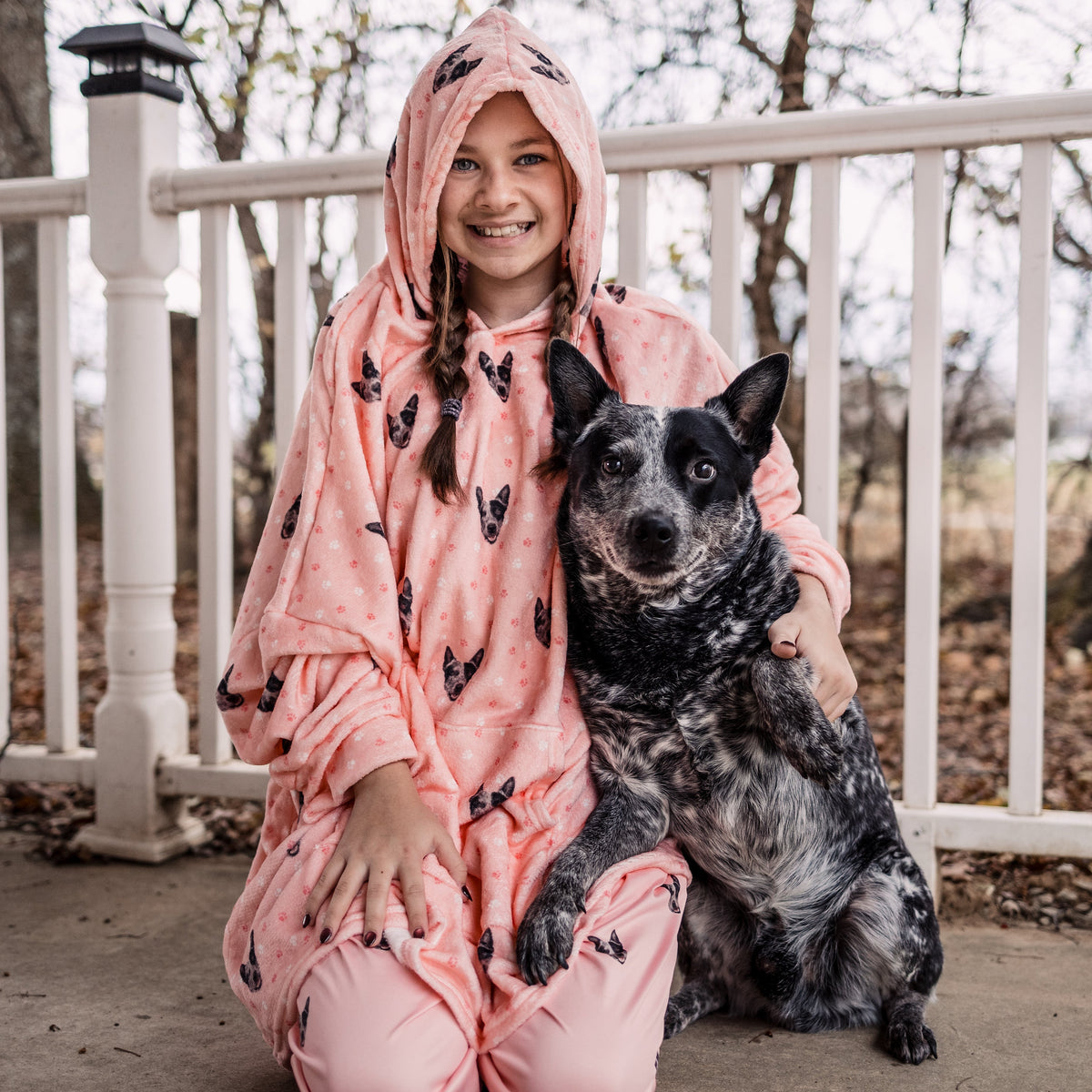 Child wearing a Light Pink Paws hooded blanket sitting on a porch beside a black and gray dog.