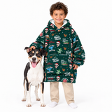 A young boy wearing a festive green Christmas-themed hooded sweatshirt stands next to a black, white, and tan dog on a solid white background. The sweatshirt features a holiday print with text like 'Dad, Bring Home All Your Christmas' and 'Santa's Gotcha'.