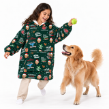 A child wearing a green Christmas-themed hooded sweatshirt with cartoon faces and text prints, playing with a golden retriever dog on a white studio background while holding a green ball.