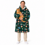 A man wearing a dark green Christmas-themed hooded blanket robe with a festive print, holding an orange tabby cat against his chest, standing against a solid white background.