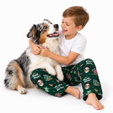 A young boy in festive green Christmas pajamas hugs a blue merle dog on a clean white studio background. The boy's pajamas feature Santa Claus, reindeer, and holiday text prints.
