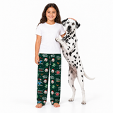 A young girl stands barefoot next to a Dalmatian dog on a white studio background, wearing dark green Christmas pajama pants with a dog-themed print and a plain white T-shirt.