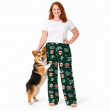 A woman wearing dark green Christmas-themed pajama pants with a corgi standing beside her on a plain white studio background. The pajama pants have an allover print of Santa hats, Christmas trees, ornaments, and the text 'Dad, come inside! It's Christmas!' and 'Dad, I love you more than Christmas!'.