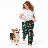 A woman wearing dark green Christmas-themed pajama pants with Santa graphics and a white t-shirt is walking barefoot next to a corgi on a white studio background.