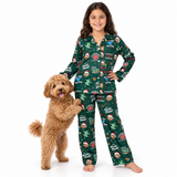 A young girl stands barefoot on a white studio background wearing a green Christmas-themed pajama set with a dog. The pajamas have an all-over festive print with Santa, snowman, Christmas tree, ornaments, and holiday text including 'DAD! CHRISTMAS!'.