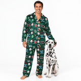 A man wearing a green Christmas-themed pajama set stands barefoot next to a Dalmatian dog on a white studio background. The pajamas feature a festive print with Santa, snowman, Christmas tree, and holiday text motifs.