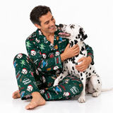 A man and a Dalmatian dog wearing matching green Christmas-themed pajamas with Dalmatian prints and holiday text, sitting together on a white studio background.