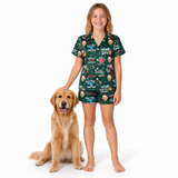 A young girl stands barefoot next to a golden retriever dog on a white studio background, wearing a dark green short-sleeve pajama set with a cartoon dog print and the words 'Don't Be A Dog'.