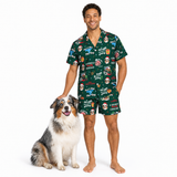 A man wearing a dark green Christmas-themed pajama set stands barefoot next to a fluffy Australian Shepherd dog on a clean white background. The pajama set features a short-sleeve button-up shirt and matching shorts with a festive print of Santa Claus, Christmas trees, ornaments, and holiday quotes.