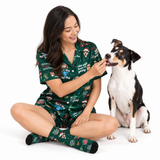 A woman wearing a green Christmas-themed pajama set with a dog sitting beside her on a white studio background. The pajamas and matching socks feature holiday illustrations and text including 'Merry Christmas,' 'The gift is on the table,' and 'The Ting.