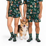 A man and a woman wearing matching dark green Christmas-themed pajama sets with a corgi dog standing between them, all photographed against a clean white studio background. The pajamas and socks feature holiday illustrations and text including 'Dad's Christmas', 'The Gift of the Christmas Tree', and 'Merry Christmas'.