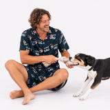 Man wearing an I Love Dad dog-print pajama short set sitting on the floor and playing tug with a black and white dog.