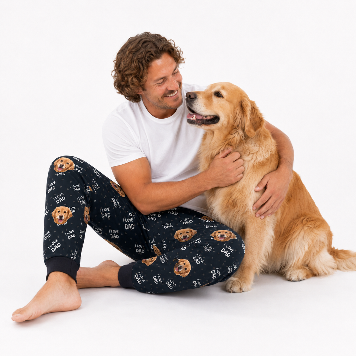 Man in black I Love Dad dog-print sweatpants sitting on floor hugging a golden retriever, white background