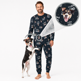 A man wearing a matching black long-sleeve pajama set with a repeating 'I LOVE DAD' text pattern and small dog illustrations, standing barefoot on a white studio background while a black-and-white dog rests on his front. A circular inset at the top right shows a close-up of the pajama print design.