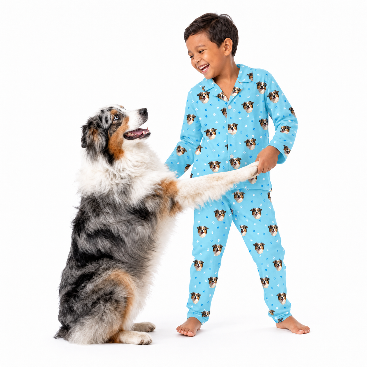 Young boy wearing a light blue paws print pajama set while smiling and holding the front paw of a happy Australian Shepherd sitting beside him.