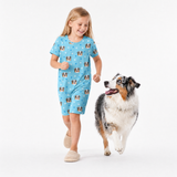 Young girl wearing a light blue paws print sleep shirt while walking in slippers beside a happy Australian Shepherd looking up at her.