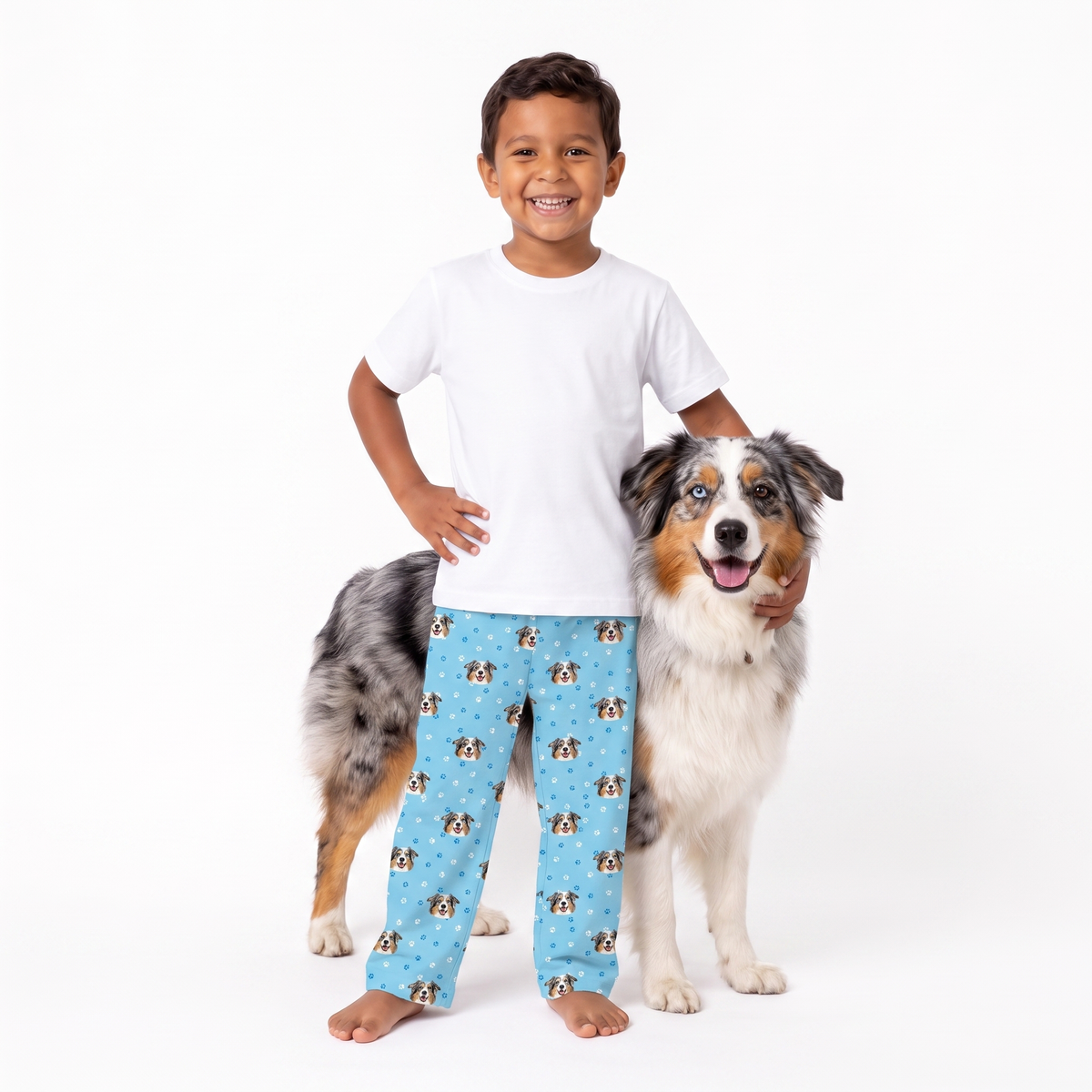 A smiling young boy stands barefoot next to a large Australian Shepherd dog on a white studio background, wearing a white t-shirt and light blue pajama pants with a repeating dog print.
