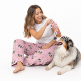 Woman wearing pink "I Love Mom" pet-print lounge pants with a white T-shirt, sitting on the floor and holding a pink plush toy above a happy Australian Shepherd lying beside her.