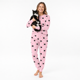 Woman wearing pink jogger pajamas with black cat patterns, holding a black and white cat on a white background