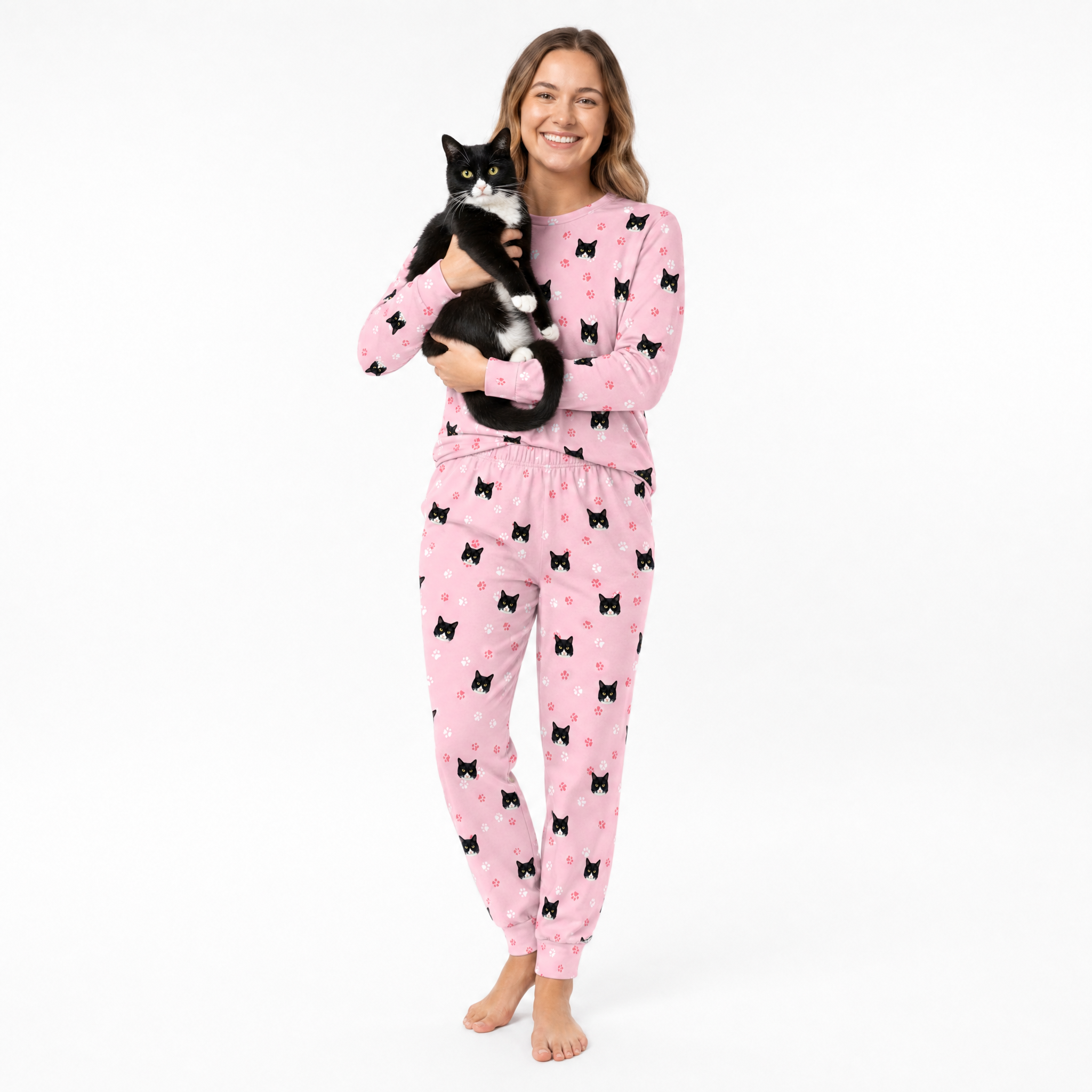 Woman wearing pink jogger pajamas with black cat patterns, holding a black and white cat on a white background