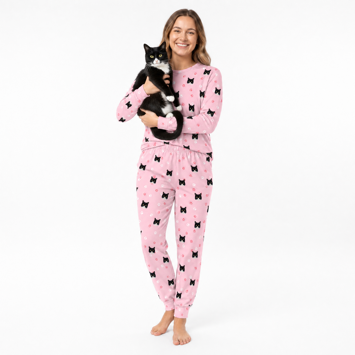 Woman wearing pink jogger pajamas with black cat patterns, holding a black and white cat on a white background