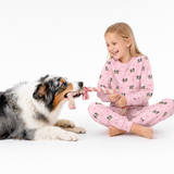 Child wearing a Pink Dream dog-print pajama set sitting on the floor and playing tug with a gray and white dog using a rope toy.