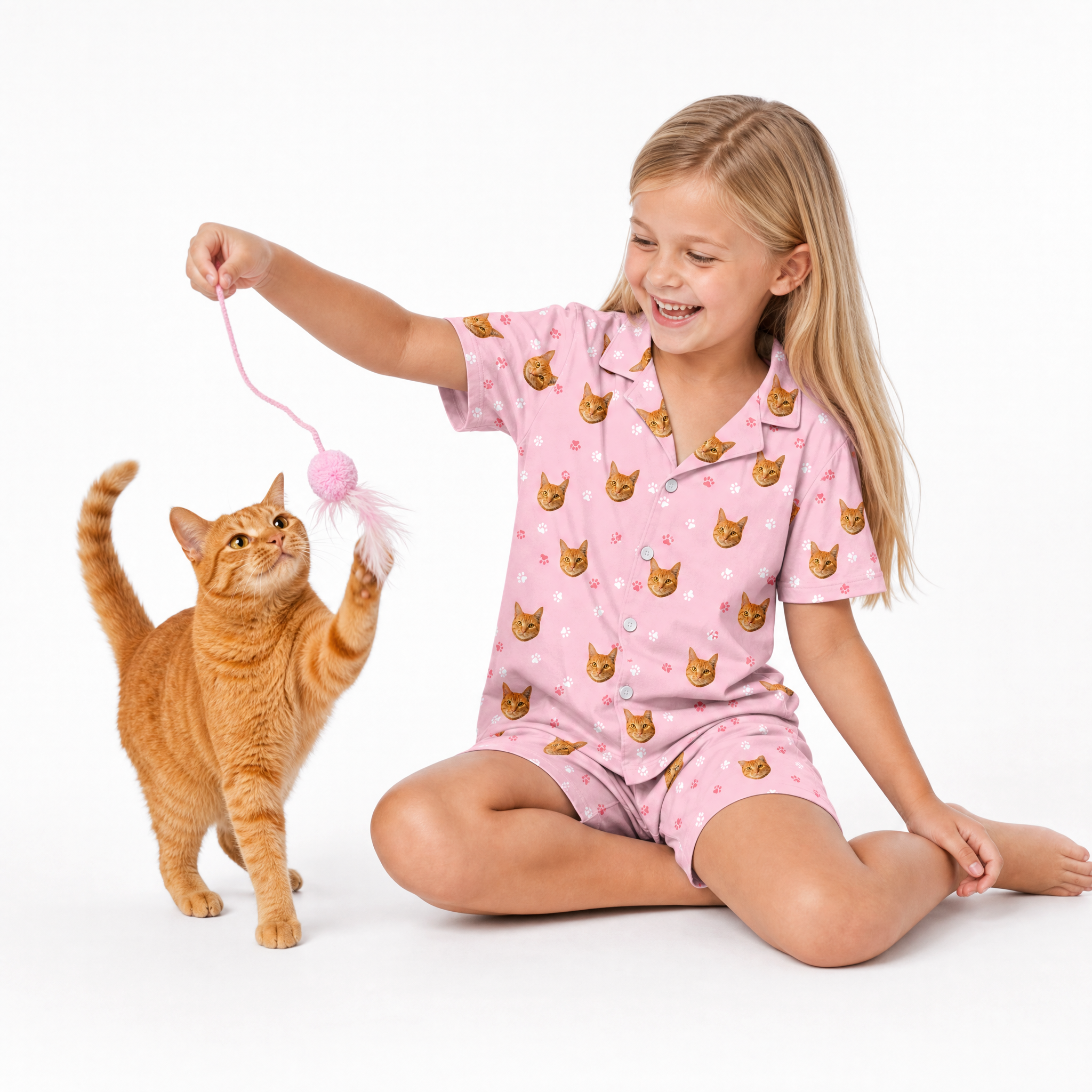 Child wearing a light pink cat-print Light pink paws pajama short set sitting on the floor and playing with an orange cat using a pink toy.