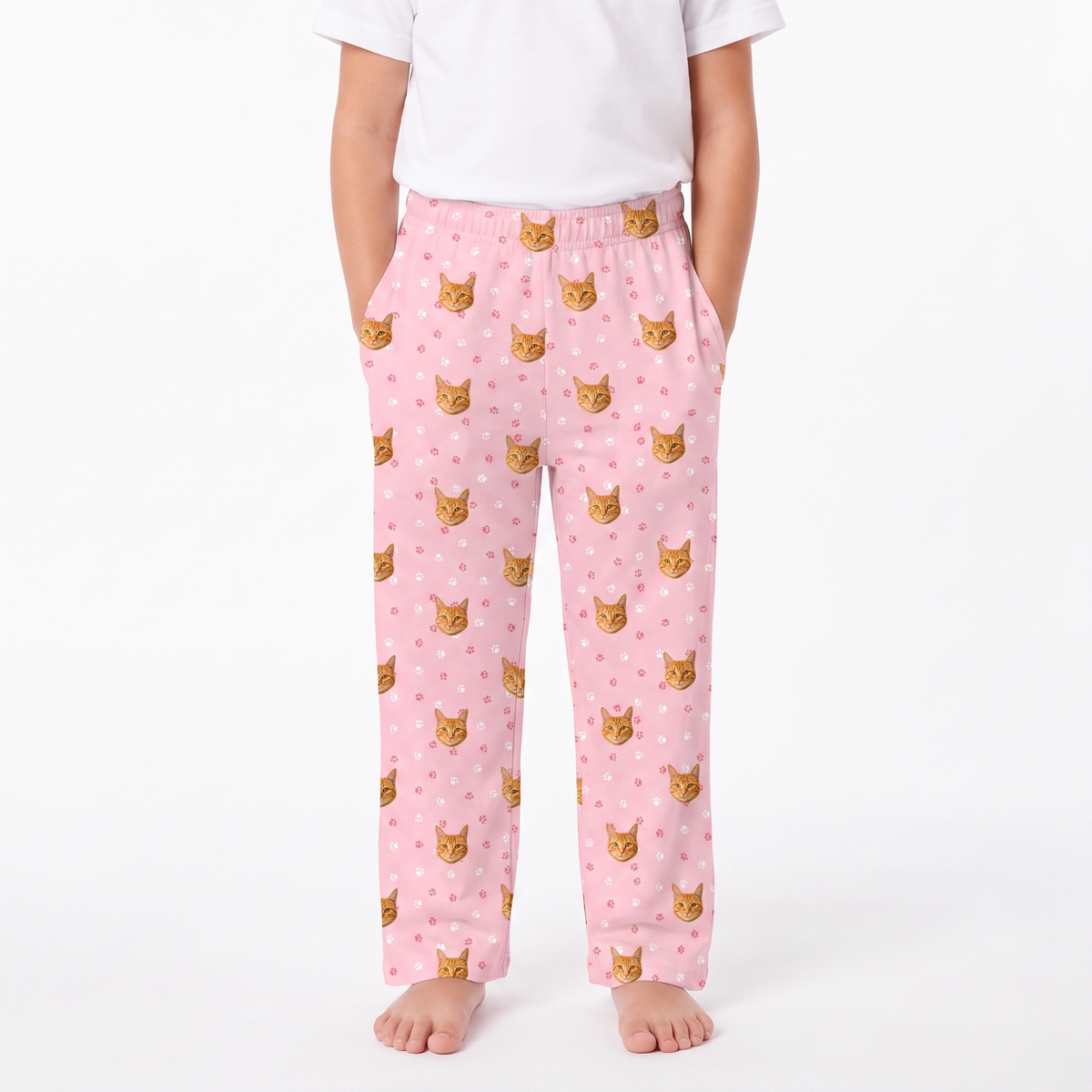 Light Pink children's pajama pants with an all-over print of orange tabby cat faces and small pink hearts, shown on a child wearing a white t-shirt against a plain white background.