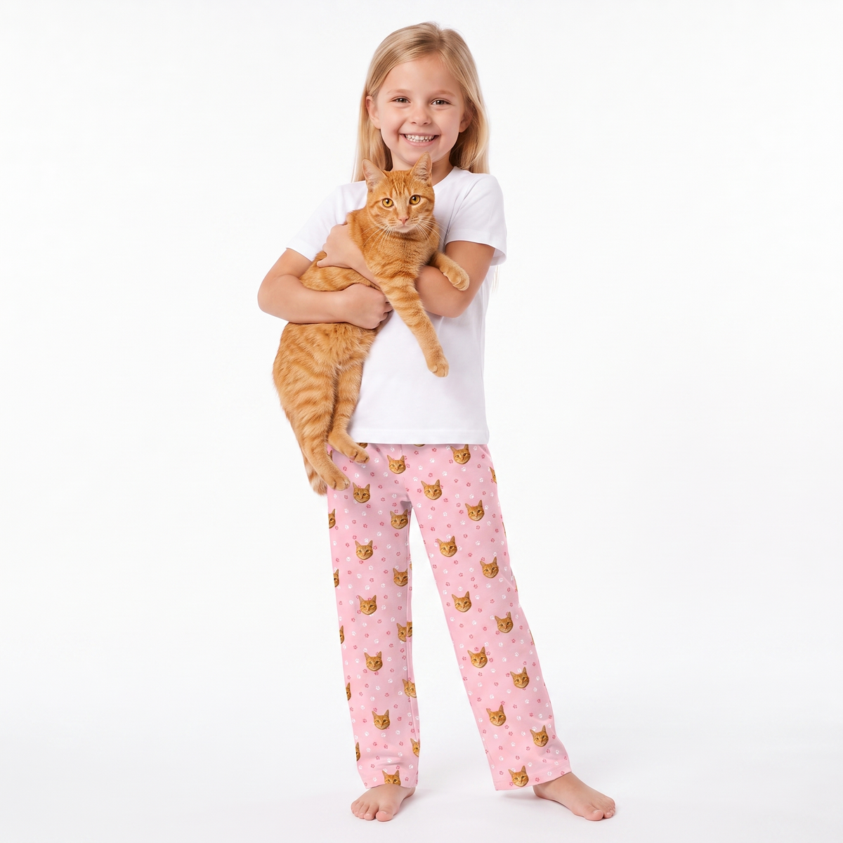 A young girl with blonde hair stands barefoot against a white studio background, holding an orange tabby cat. She is wearing a white short-sleeve t-shirt and light pink pajama pants with a repeating cat-face pattern.