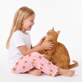 A young girl with long blonde hair sits on a white floor, smiling and petting an orange tabby cat. She is wearing a white t-shirt and light pink pajama pants with a repeating pattern of sleeping cats.