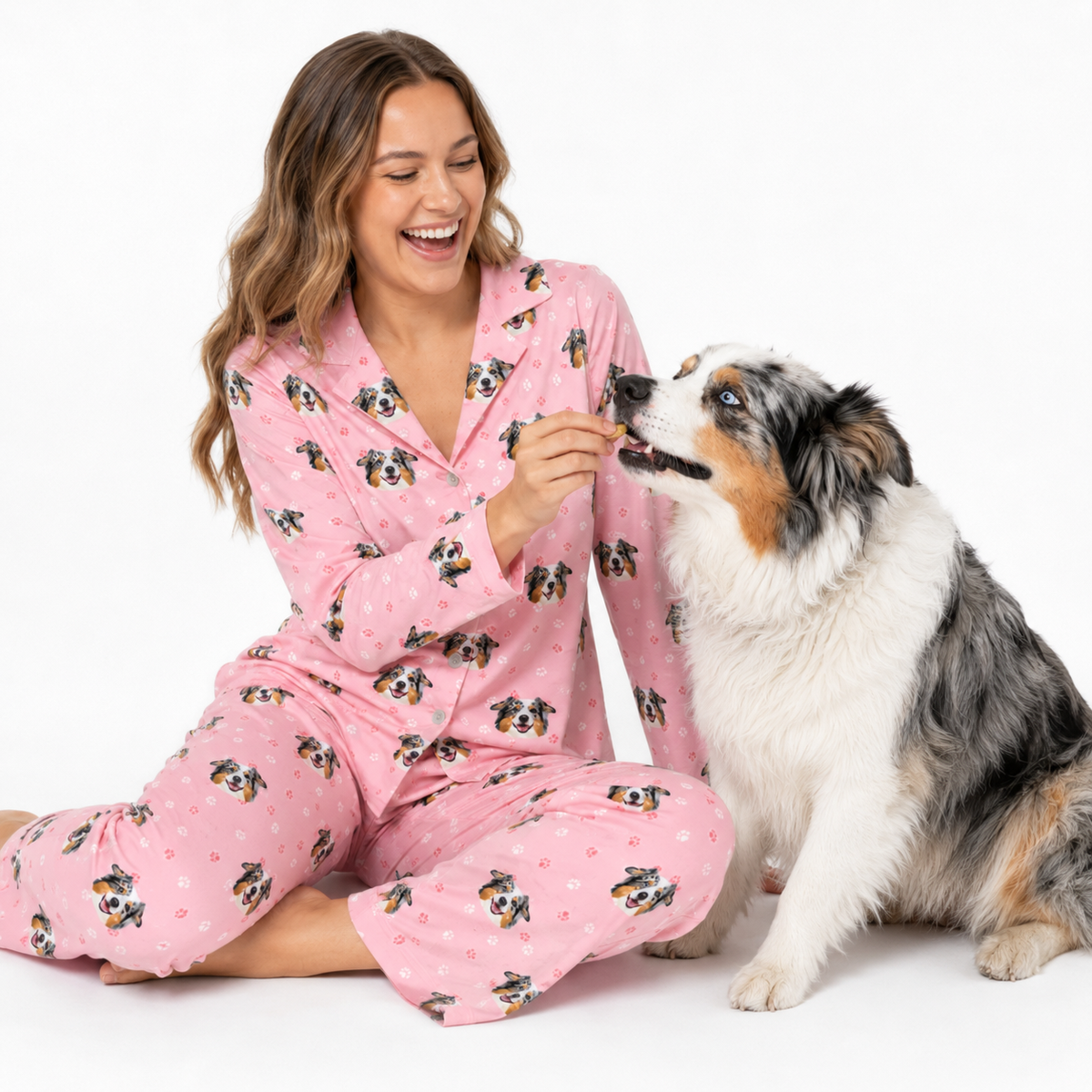 Woman wearing a light pink paws dog-print pajama set sitting on the floor and giving a treat to a gray and white dog.