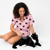 Woman wearing a Light Pink Paws cat-print pajama set sitting on the floor and brushing a black and white cat.