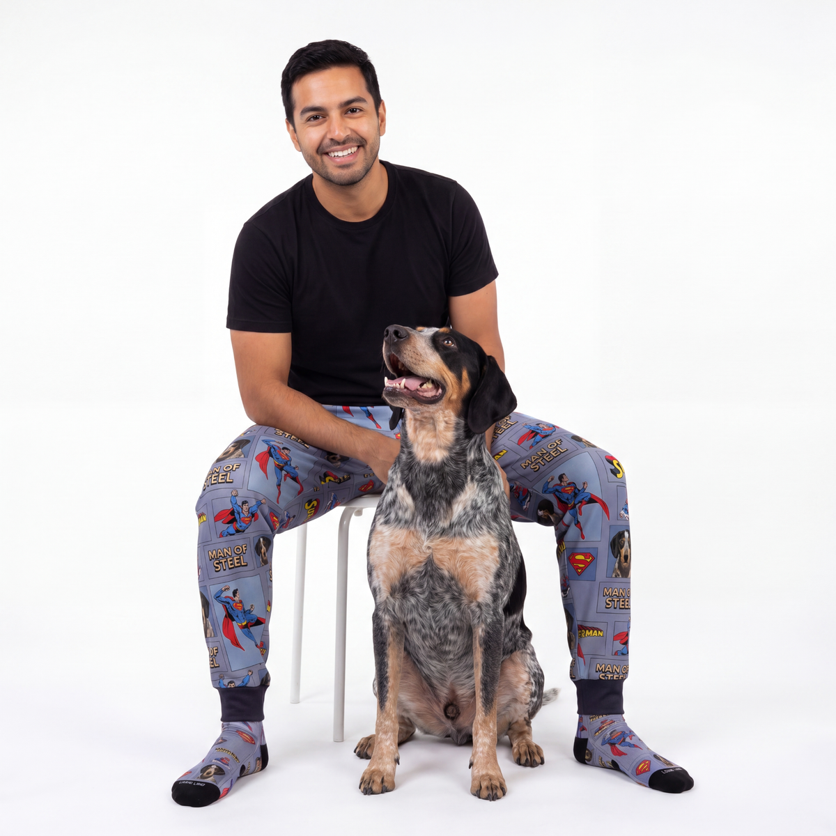 Man wearing superman pajama sweatpants sitting on a chair with a dog.