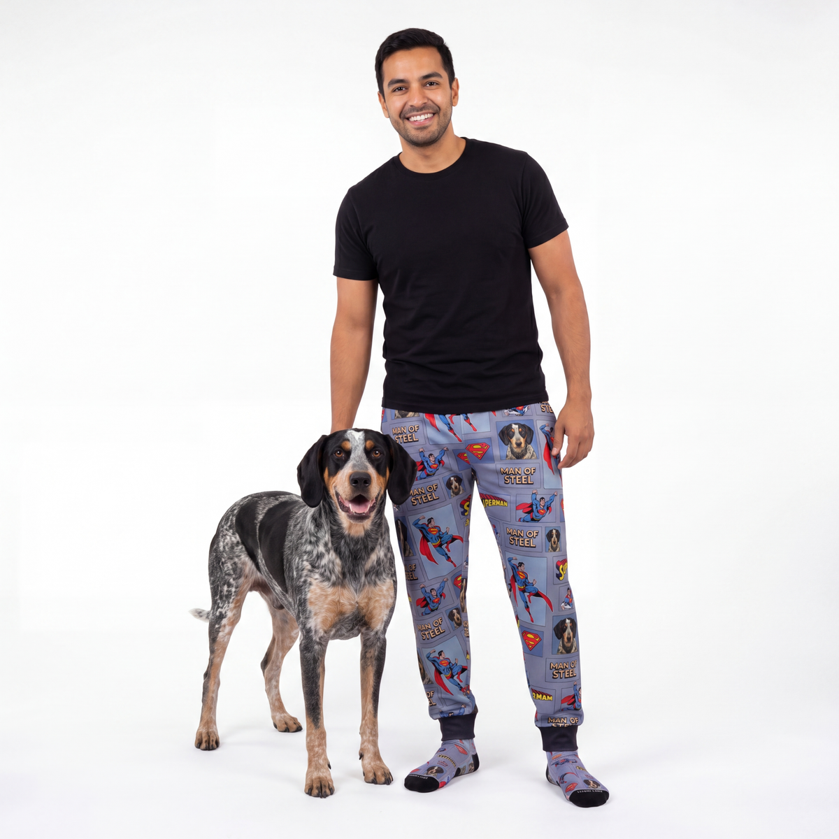 Man wearing superman pajama sweatpants standing next to a dog on a white background