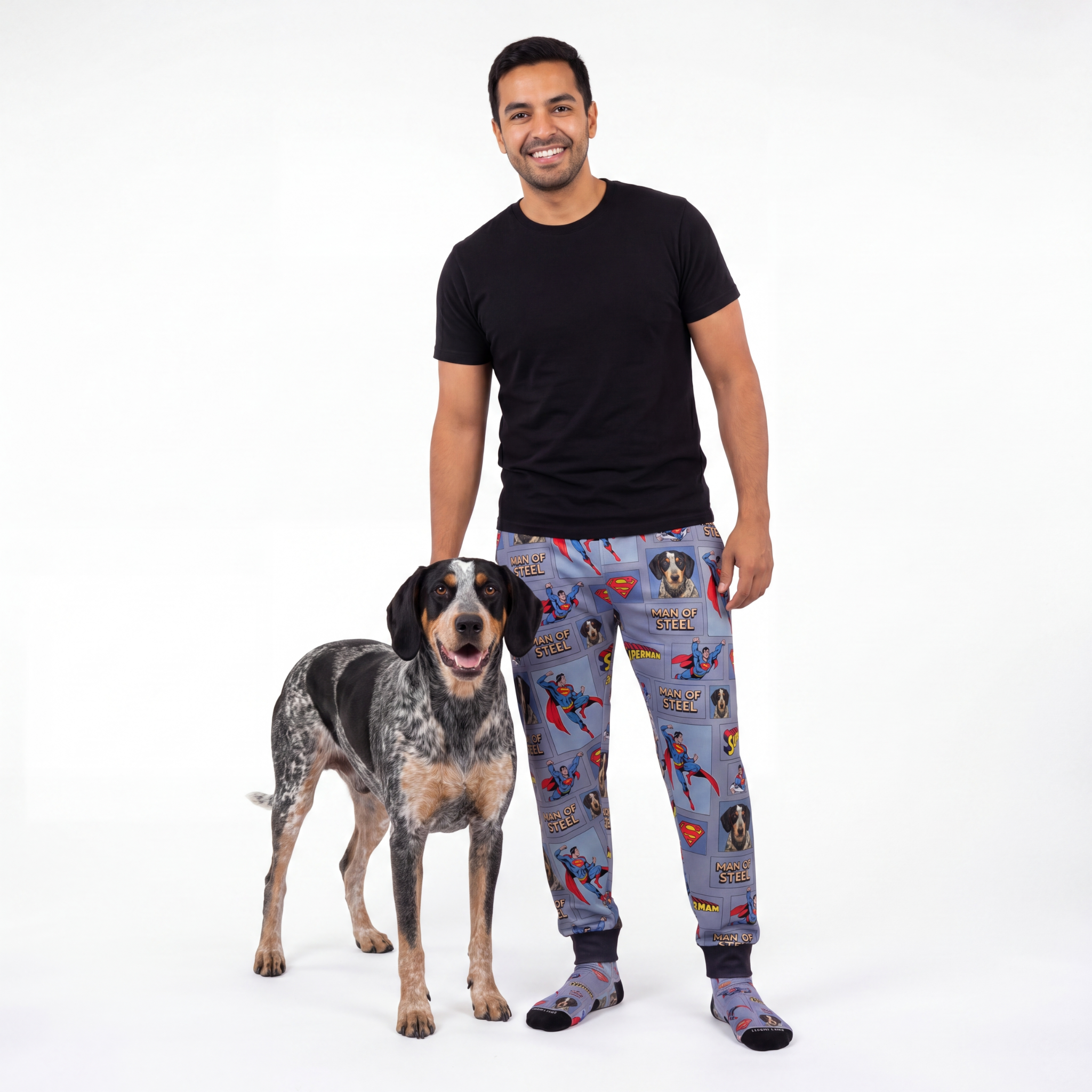 Man wearing superman pajama sweatpants standing next to a dog on a white background