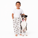 Child wearing pajama pants with dog pattern standing next to a dog on a white background