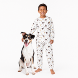 Child wearing a white jogger pajama set with dog pattern standing next to a dog on a white background