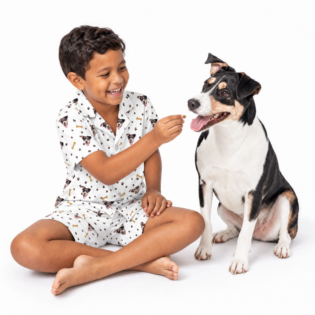 Child in white pajamas with dog and bone prints sitting next to a dog on a white background