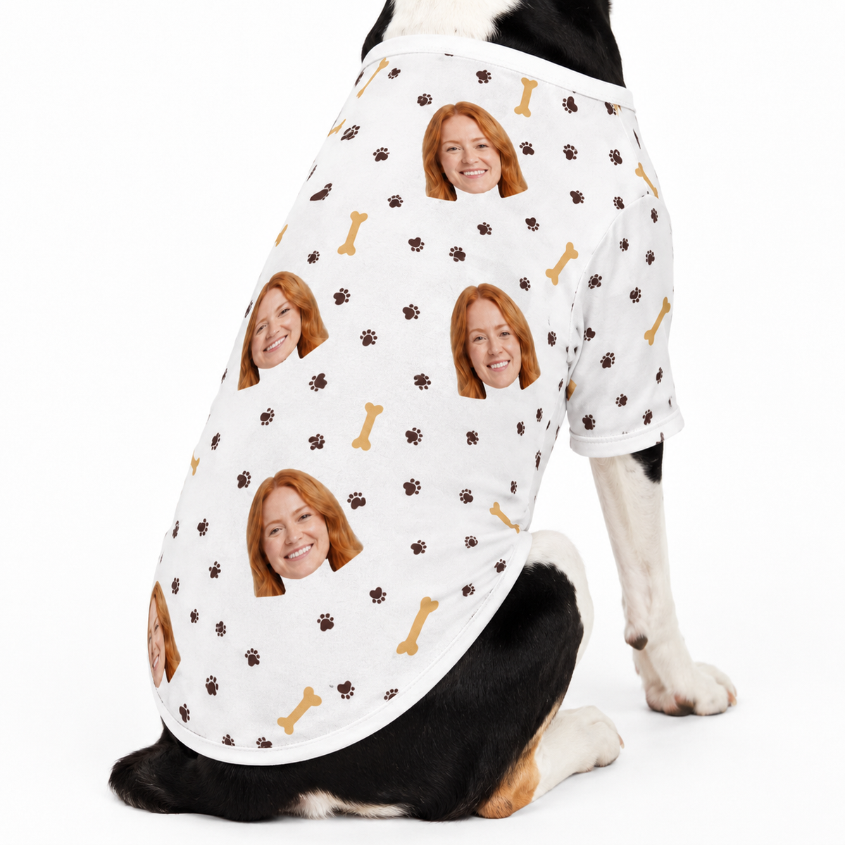 Dog wearing a white custom dog shirt with a repeating face, paw print, and bone pattern on a white studio background.