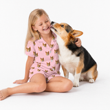 Child wearing a Pink Dream dog-print pajama set sitting beside a corgi that is licking her cheek.