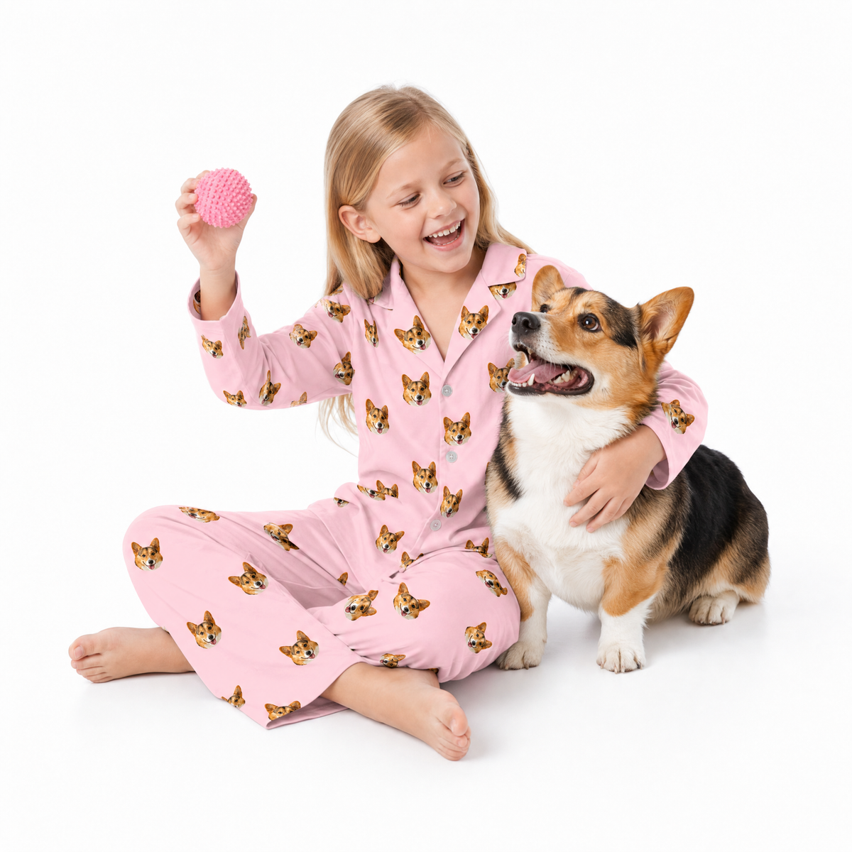 Child in pink pajamas with dog pattern sitting on a white background, holding a pink ball and petting a corgi.
