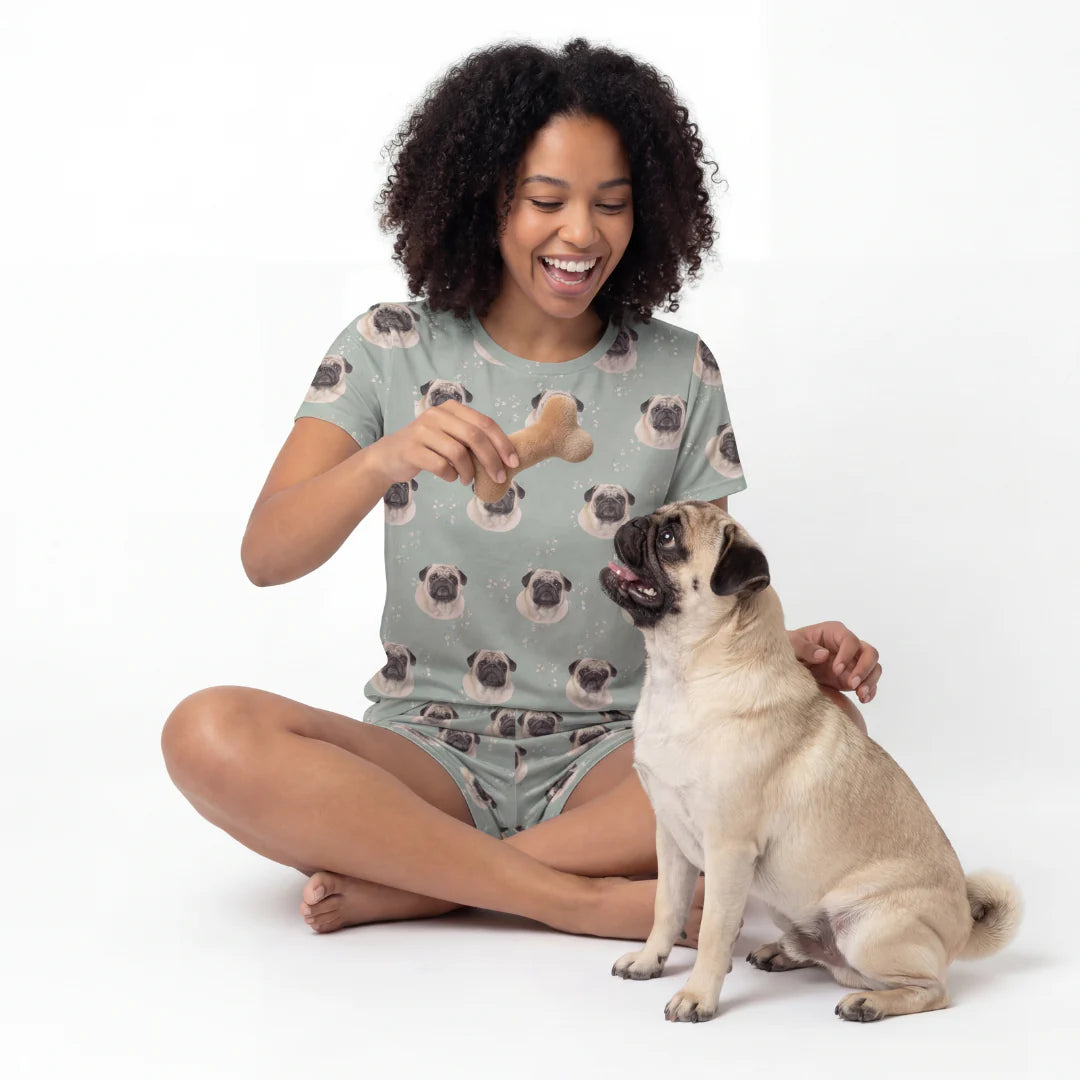 Woman in pug-themed pajamas holding a bone-shaped toy with a pug sitting next to her on a white background