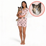 A woman stands barefoot against a white background holding a small dog, wearing a light pink pajama set printed with dog faces. A circular inset at the top right shows a close-up of the dog's face with a pink ring around it, linking the product to the pet.