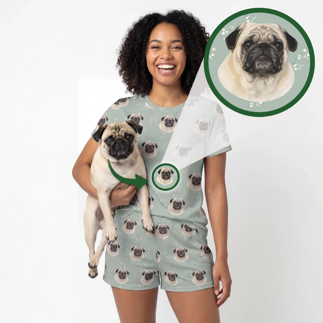 A woman wearing a matching pajama set with an all-over pug print, holding a small pug dog. The set consists of a light sage green t-shirt and shorts with a repeating pattern of the pug's face.