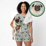 A woman wearing a matching pajama set with an all-over pug print, holding a small pug dog. The set consists of a light sage green t-shirt and shorts with a repeating pattern of the pug's face.
