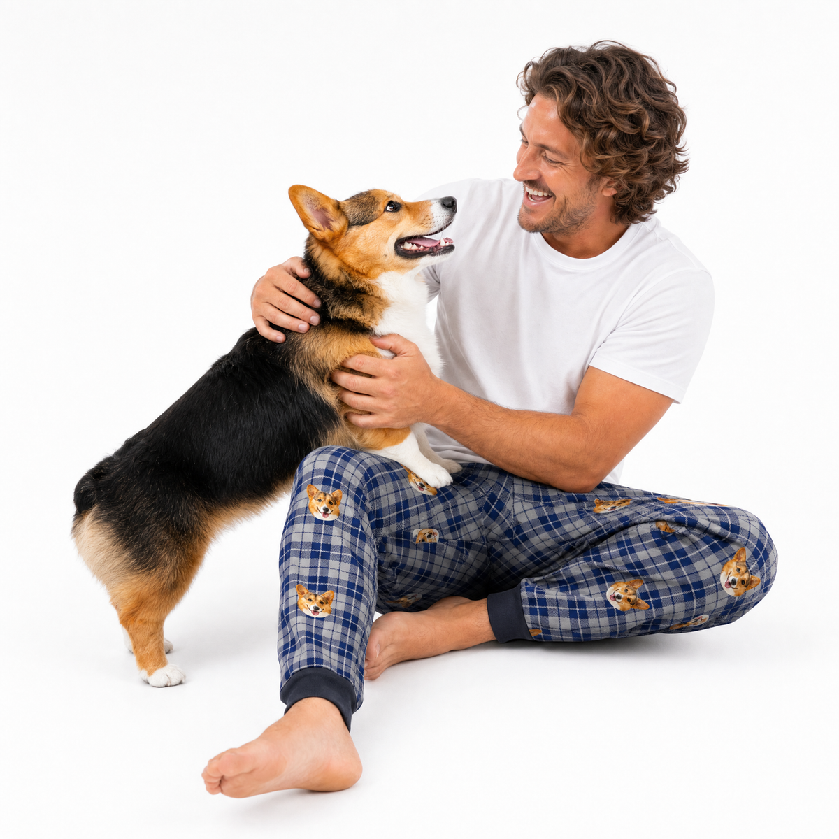 A smiling man sitting on a white studio floor with his legs crossed, wearing a white t-shirt and blue plaid sweatpants printed with corgi faces. He is holding and petting a black, tan, and white Corgi dog against a seamless white background.