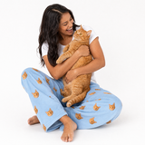 Woman wearing a white T-shirt and sky blue cat-print lounge pants, sitting cross-legged on the floor while smiling and cuddling an orange tabby cat in her arms.