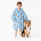Child wearing a sky blue blanket hoodie with dog pattern standing next to a corgi on a white background